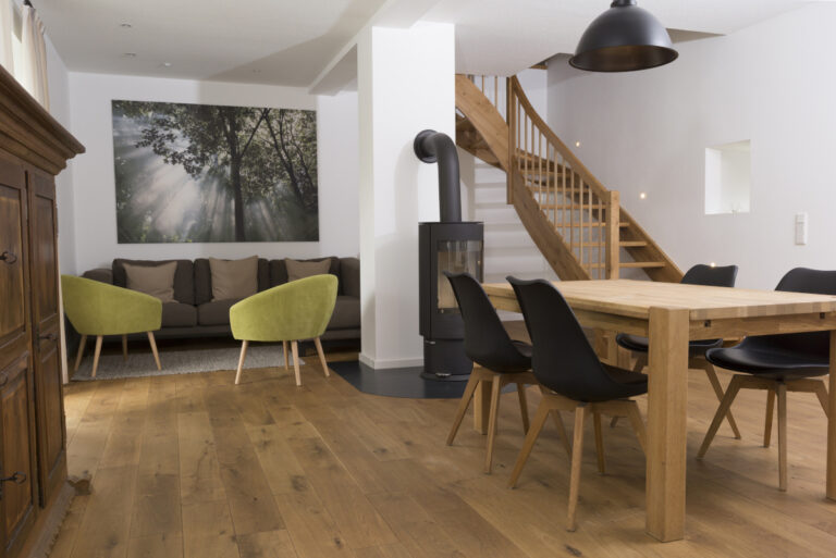 Wohn-Esszimmer mit Holzboden, Holztisch, braunem Sofa und zwei grünen Sesseln, Holzofen