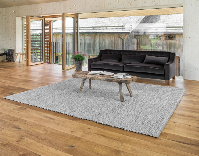 Wohnzimmer mit großer Fensterfront, schwarzes Sofa, rustikaler Couchtisch und Webteppich