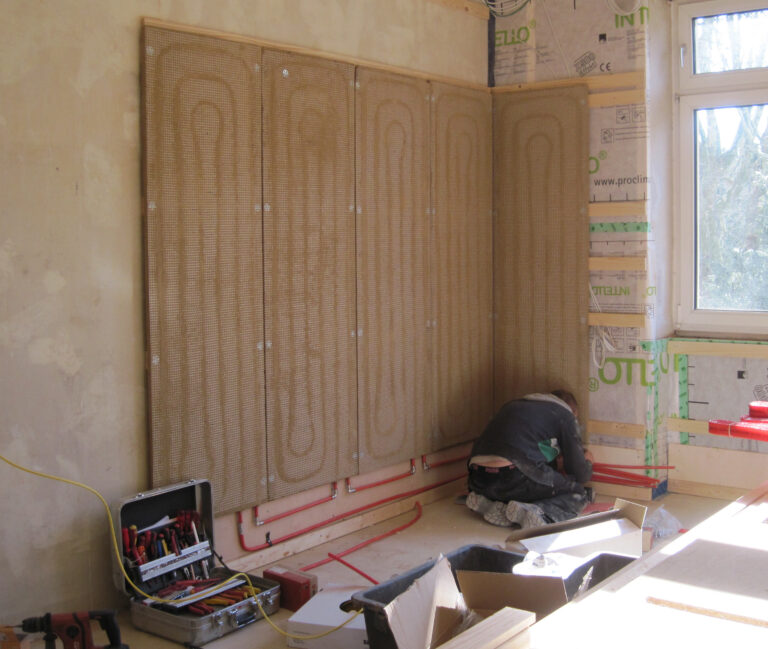 Zimmer mit rohen Wänden, an die ein Arbeiter eine Wandheizung montiert