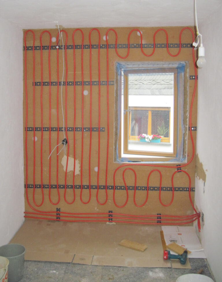 Zimmer mit rohen Wänden, von der Decke hängt Glühbirne, auf Außenwand wurden Rohre für Wandheizung angebracht