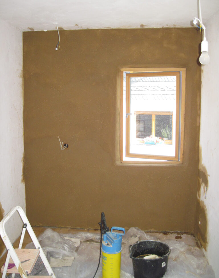 Zimmer mit rohen Wänden, von der Decke hängt Glühbirne, Außenwand mit braunem Lehm verputzt