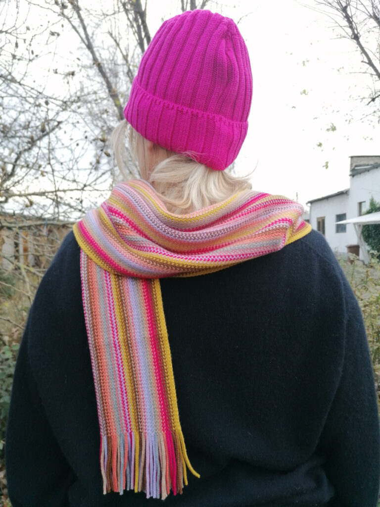Frau mit blonden Haaren trägt eine pinkfarbene Wollmütze und einen passenden bunt-gestreiften Wollschal