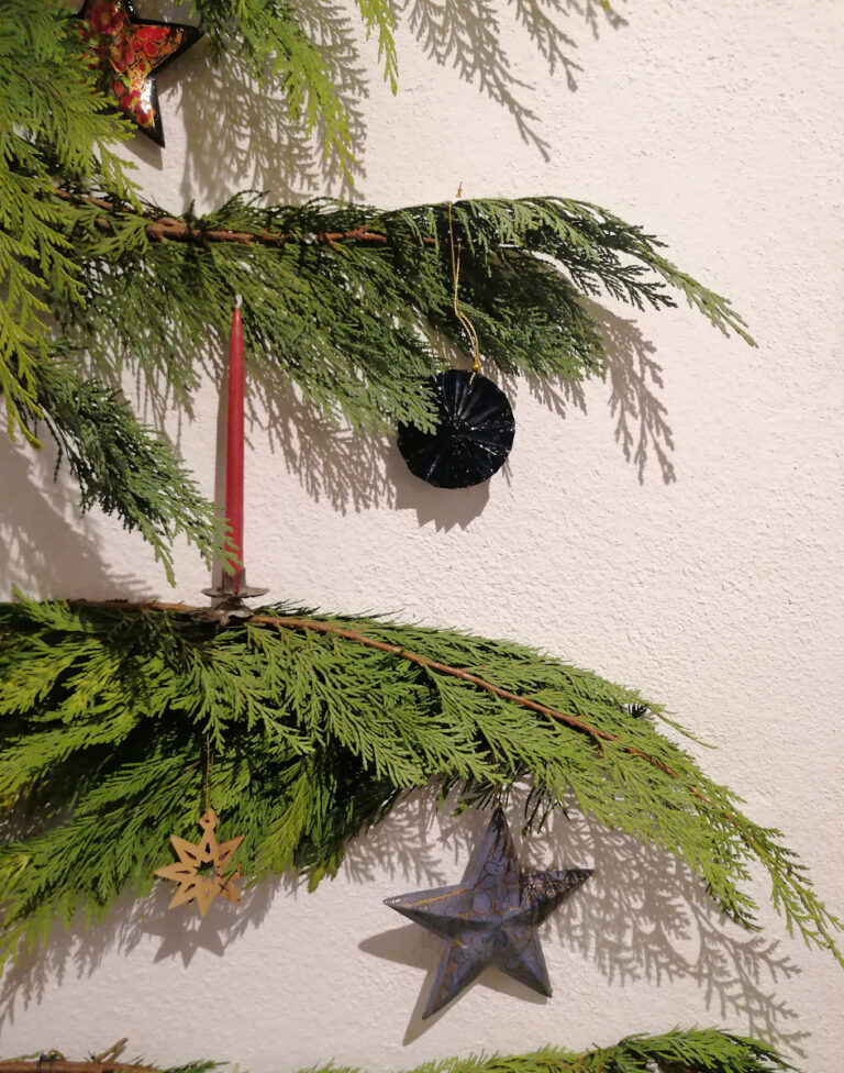 Weihnachtsornamente aus verschiedenen Materialien hängen an einem Tannenzweig vor einer weißen Wand