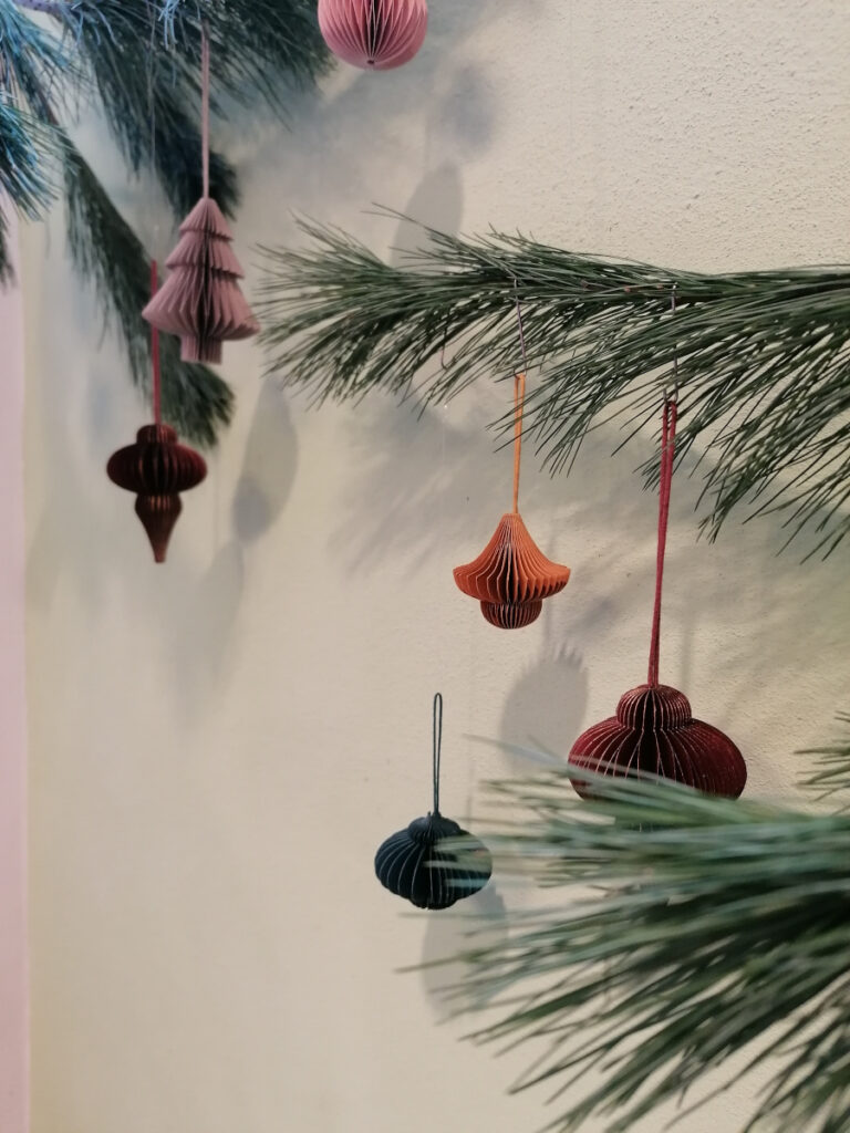 dreidimensionale Weihnachtsornamente aus Papier hängen an einem Tannenzweig vor einer weißen Wand