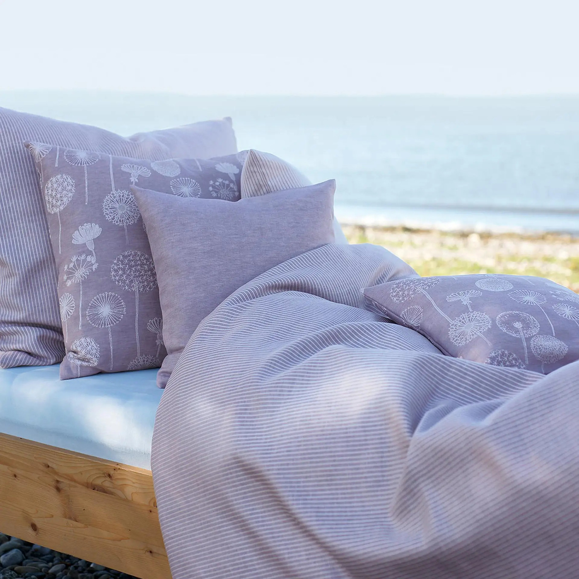 Bett, das an einem Strand steht mit fliederfarbener Bettdecke und Kissen